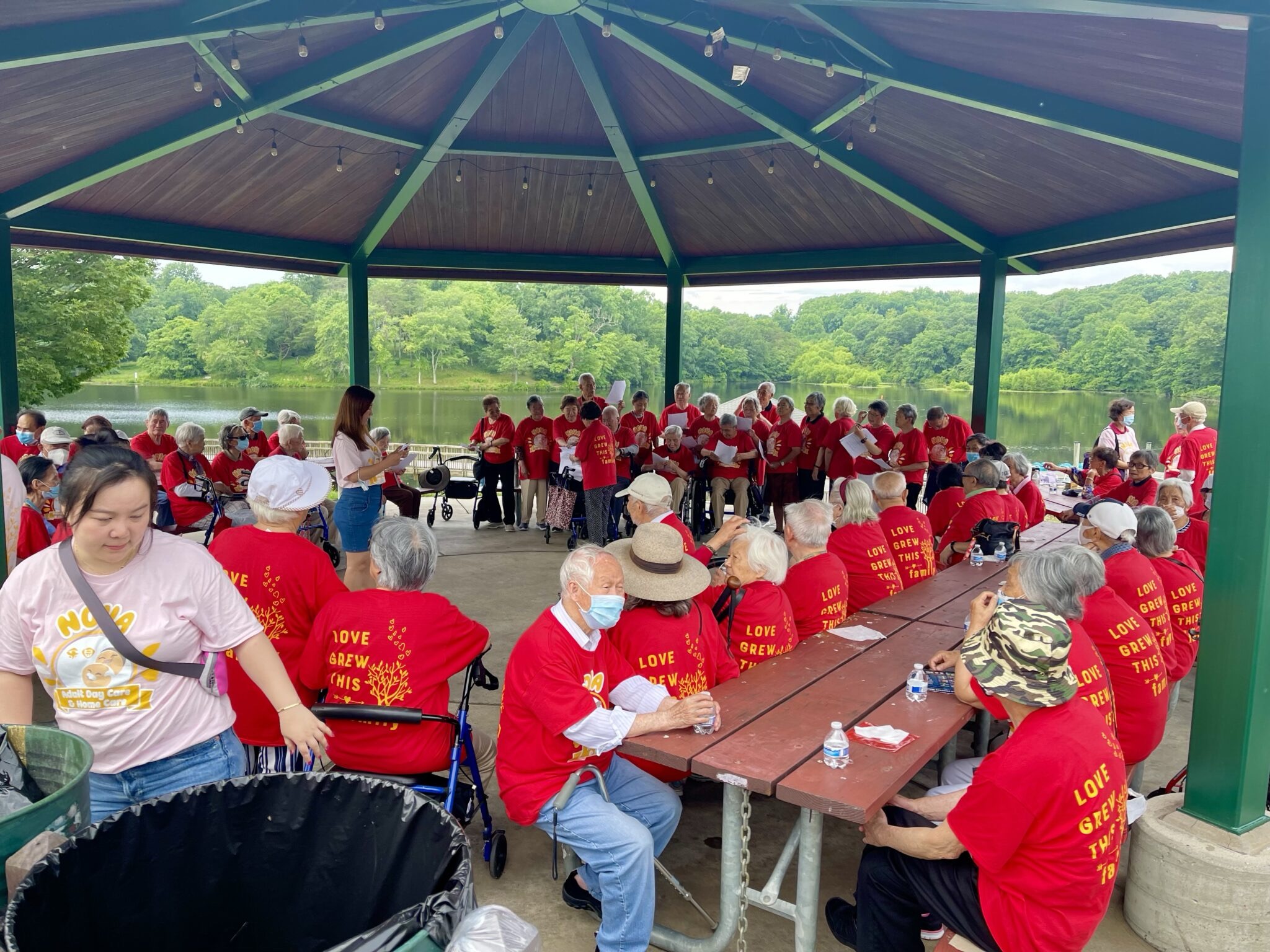 Seniors in matching red shirts singing together at a lakeside pavilion event